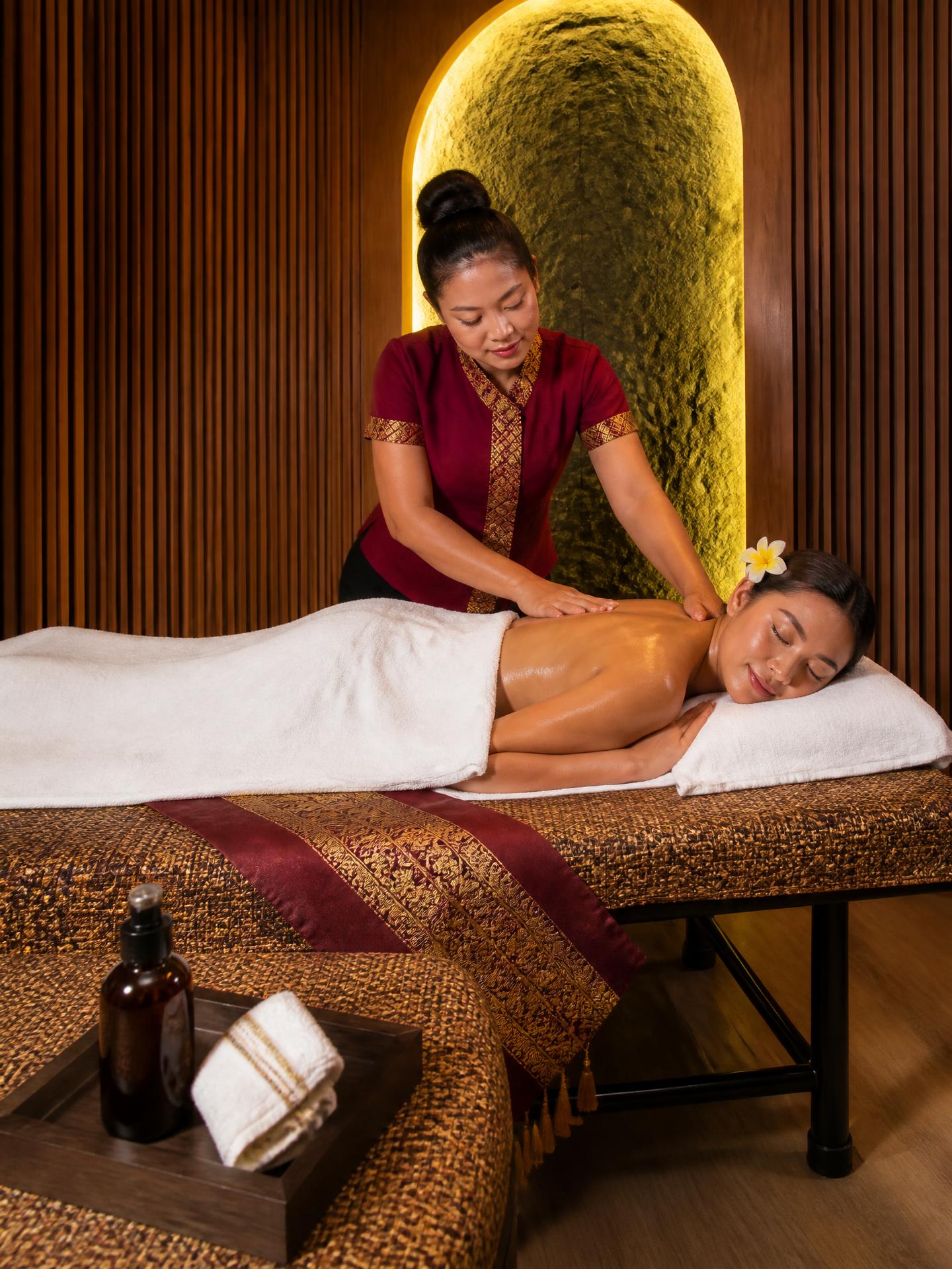 A Thai Oasis therapist working warm aromatherapy oil into a guest's shoulders on a bed dressed with a burgundy-and-gold Thai runner